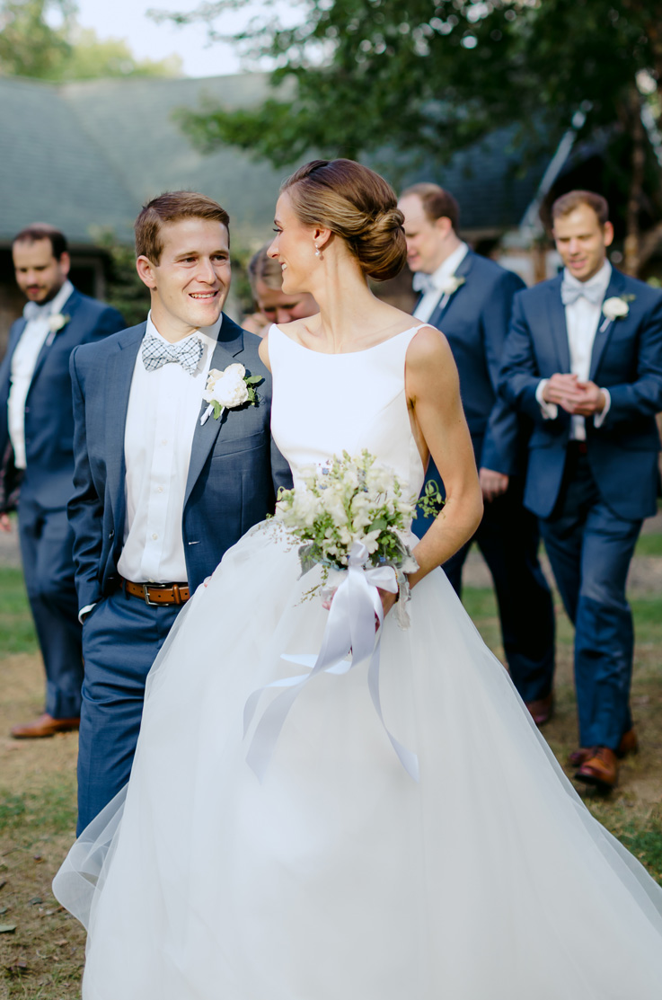 A bride in a sleeveless ball gown wedding dress, holding a bouquet, and a groom in a suit smile and walk together outside, with several groomsmen walking in the background.