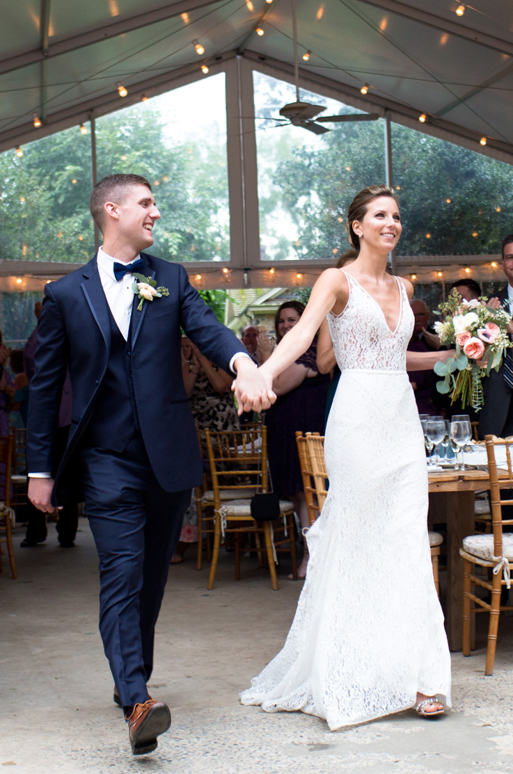 A groom in a suit is holding hands with a bride in a V-neck lace wedding dress and smiling at her, as they both walk inside an enclosed outdoor tent, with wedding guests, tables, and chairs in the background.