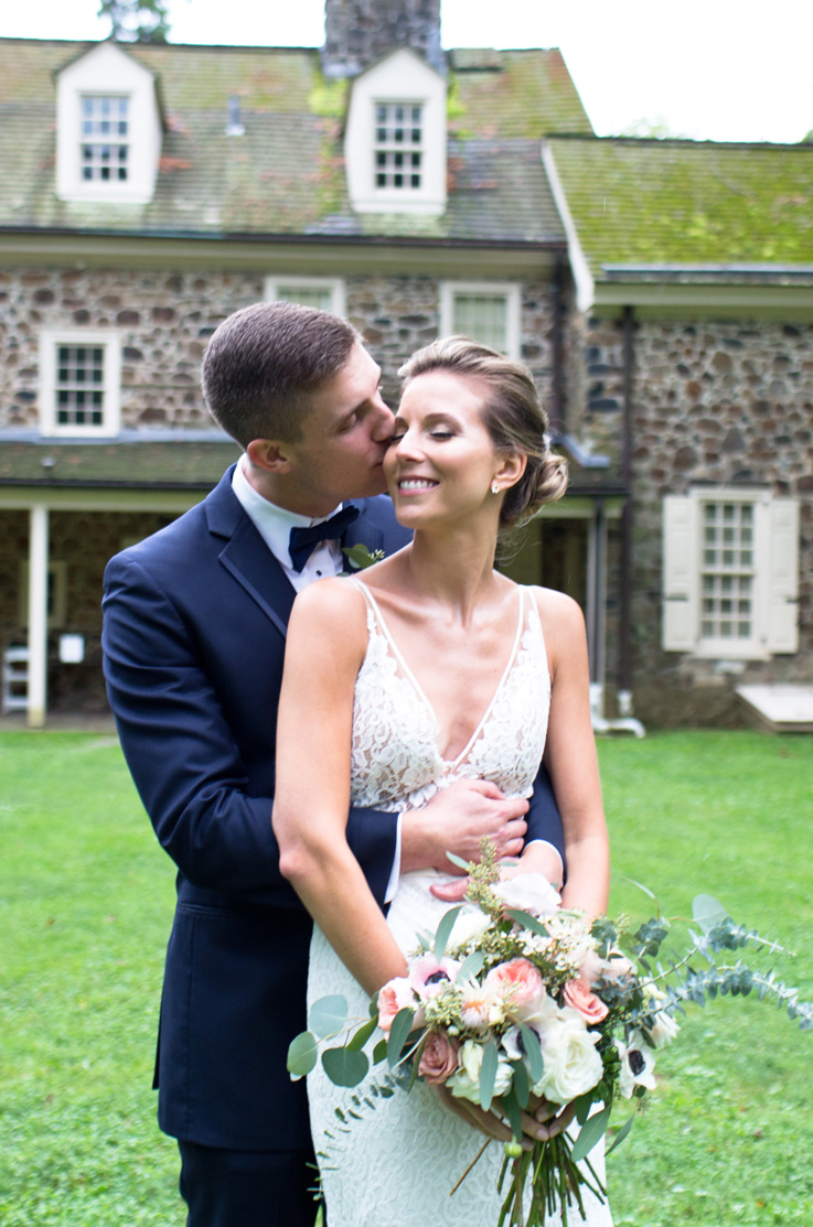 A groom in a suit stands behind a bride in a V-neck lace wedding dress and kisses her on the cheek as she smiles and holds a bouquet, they are standing outside in front of a stone house.