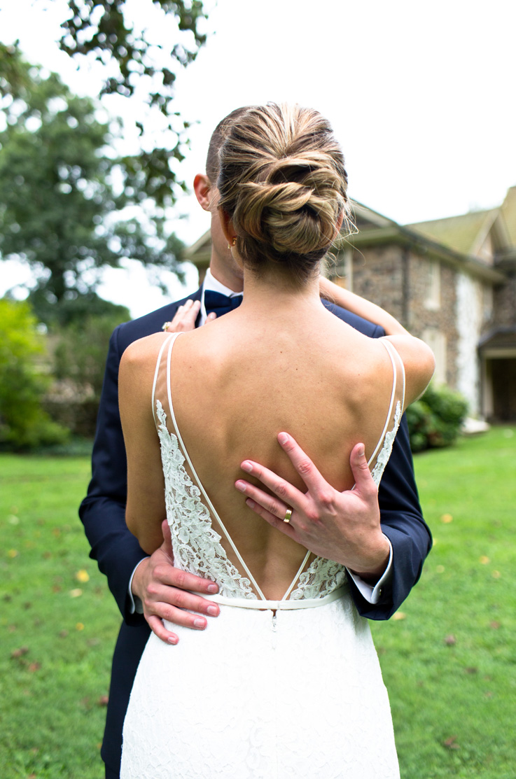 A bride in a sleeveless lace wedding dress stands in front of a groom, facing him, with the groom's arms around her, as they with stand outside in the grass with trees and a house behind them.