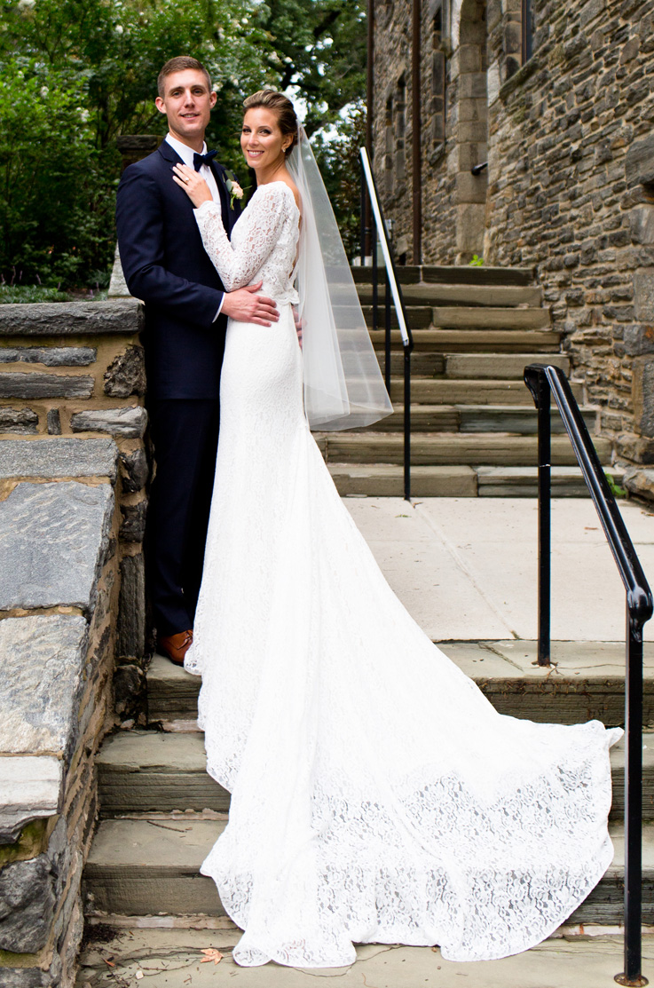 A bride in a long sleeve lace wedding dress and veil, and a groom in a suit, stand on a stone staircase outside of a stone building, with the bride's lace dress train flowing down the stairs.