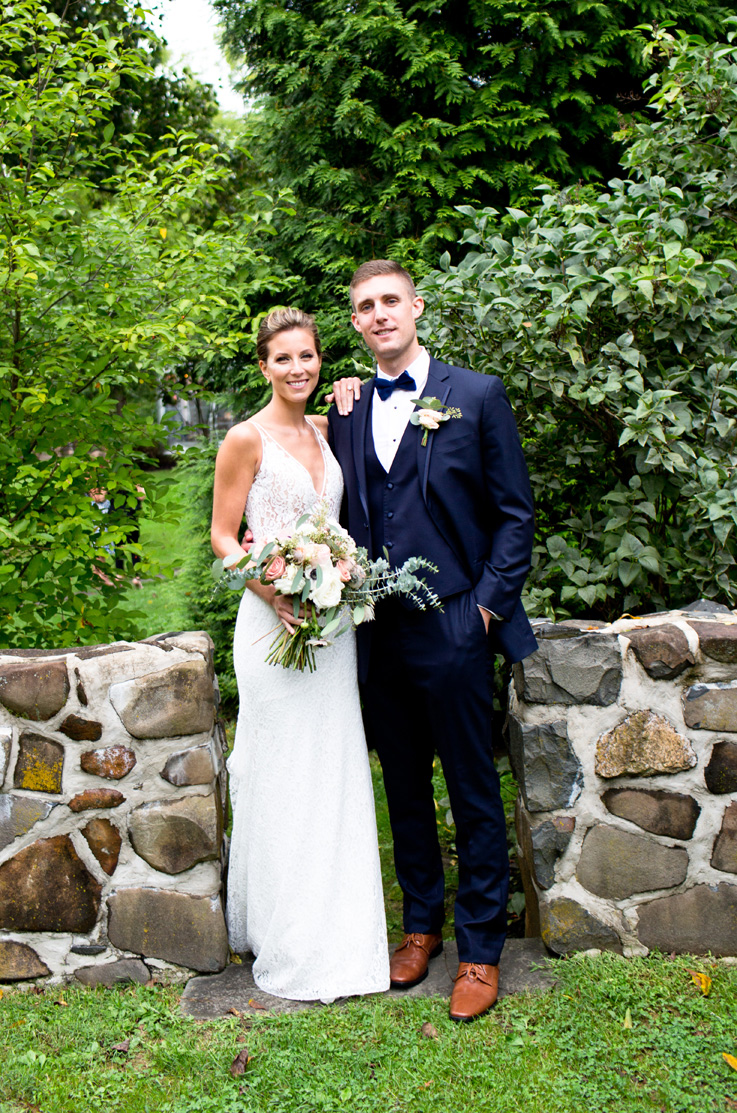 A bride in a V-neck lace wedding dress holding a bouquet is standing outside with a groom in a suit with her hand on his shoulder, both of them are smiling and are in front of green bushes and a short stone wall.