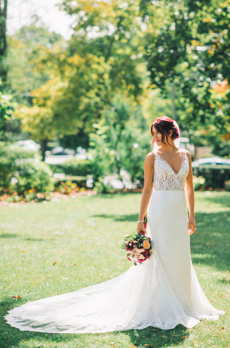 A bride in a V-neck lace wedding dress holds a bouquet and poses outside in the grass, in front of trees and a garden, with her lace train fanned out beside her.