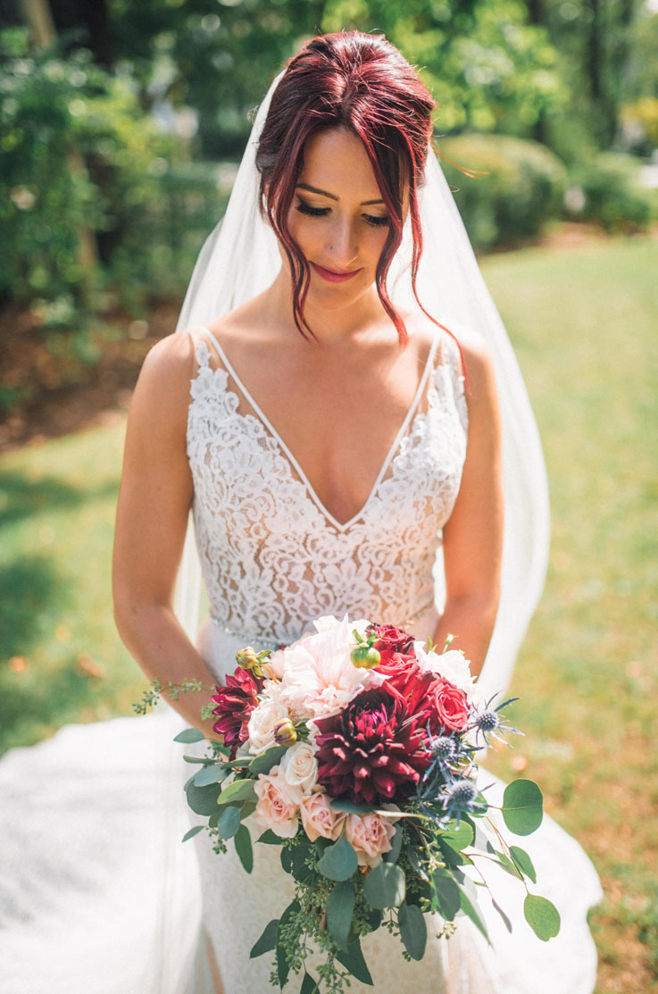A bride in a V-neck lace wedding dress and veil, holding a bouquet, stands outside in the grass with trees in the background.