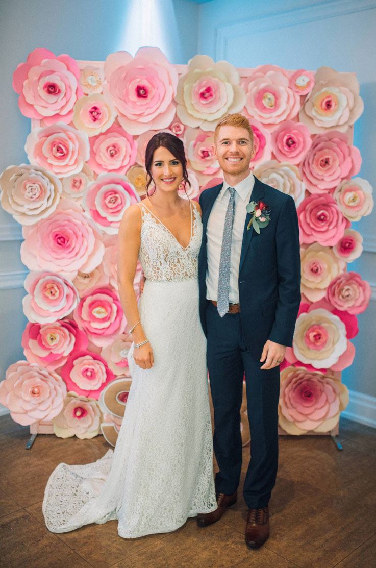 A bride in a V-neck lace wedding dress and a groom in a suit smile and pose together in front of a paper flower wall, inside a ball room.