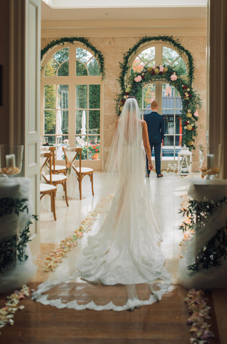 A bride in a sleeveless lace wedding dress and veil walks towards a groom in a suit in a decorated ball room, the groom is facing away and is moments away from seeing his bride for their first look.