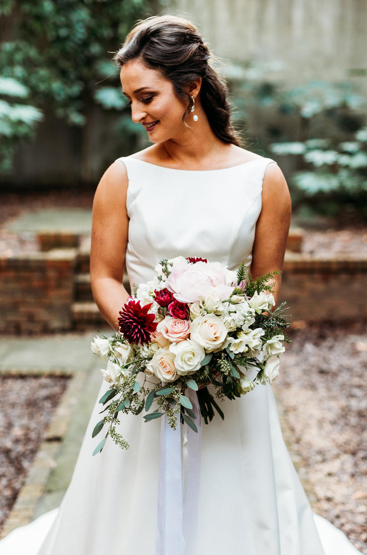 A bride in a sleeveless wedding dress, holding a bouquet, poses in a brick courtyard on a path while looking down towards her shoulder and smiling.