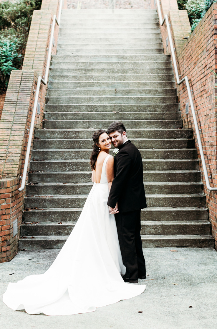 A bride in a sleeveless wedding dress and a groom in a suit stand beside each other smiling and holding hands, while standing at the bottom of a brick and concrete staircase. 