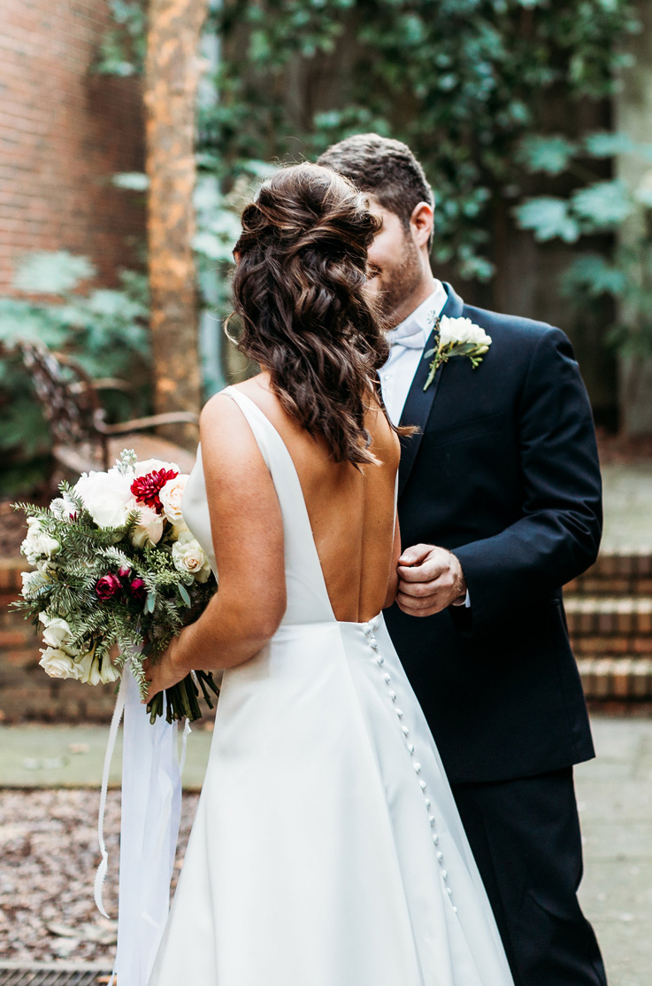 A bride in a sleeveless wedding dress, holding a bouquet, and a groom in a suit stand outside in a brick courtyard with foliage and a bench in the background.  
