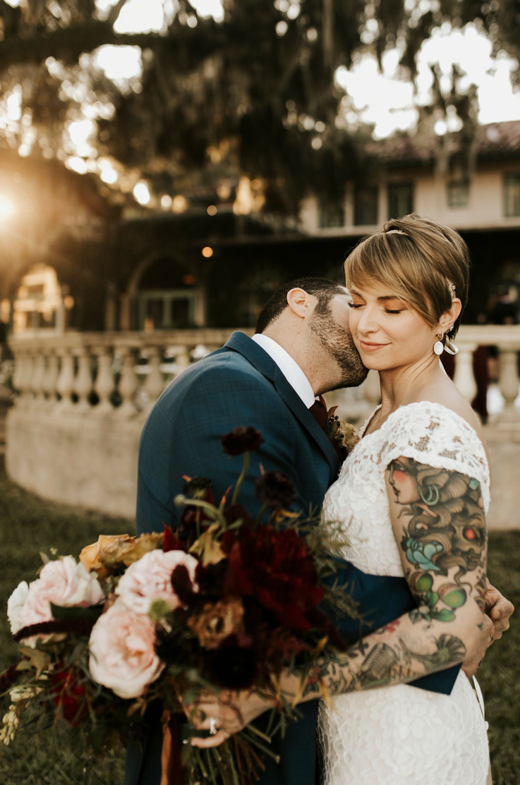 A groom in a suit holds a bride in a cap sleeve lace wedding dress and kisses her on the cheek, as they both stand outside in front of a villa.