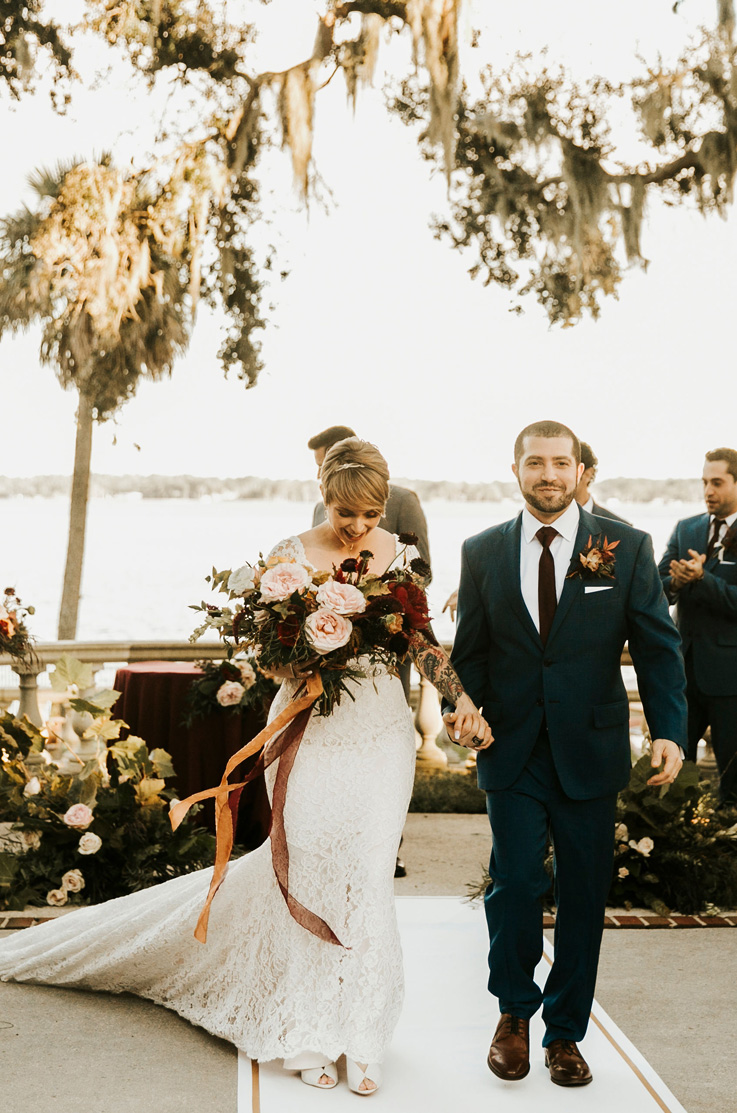A bride in a cap sleeve lace wedding dress, holding a bouquet, and a groom in a suit hold hands in front of a few groomsmen, with a lake and trees behind them.