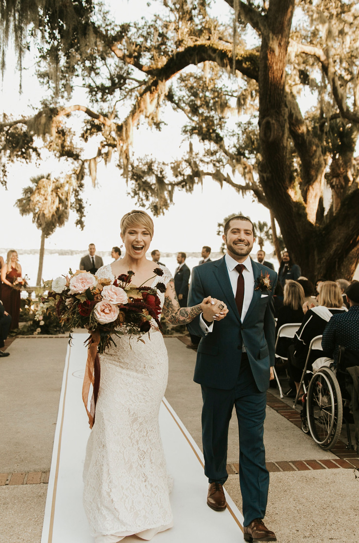 A bride in a cap sleeve lace wedding dress, holding a bouquet, and a groom in a suit, smile and hold hands while walking down the aisle outside, with wedding guests behind them.