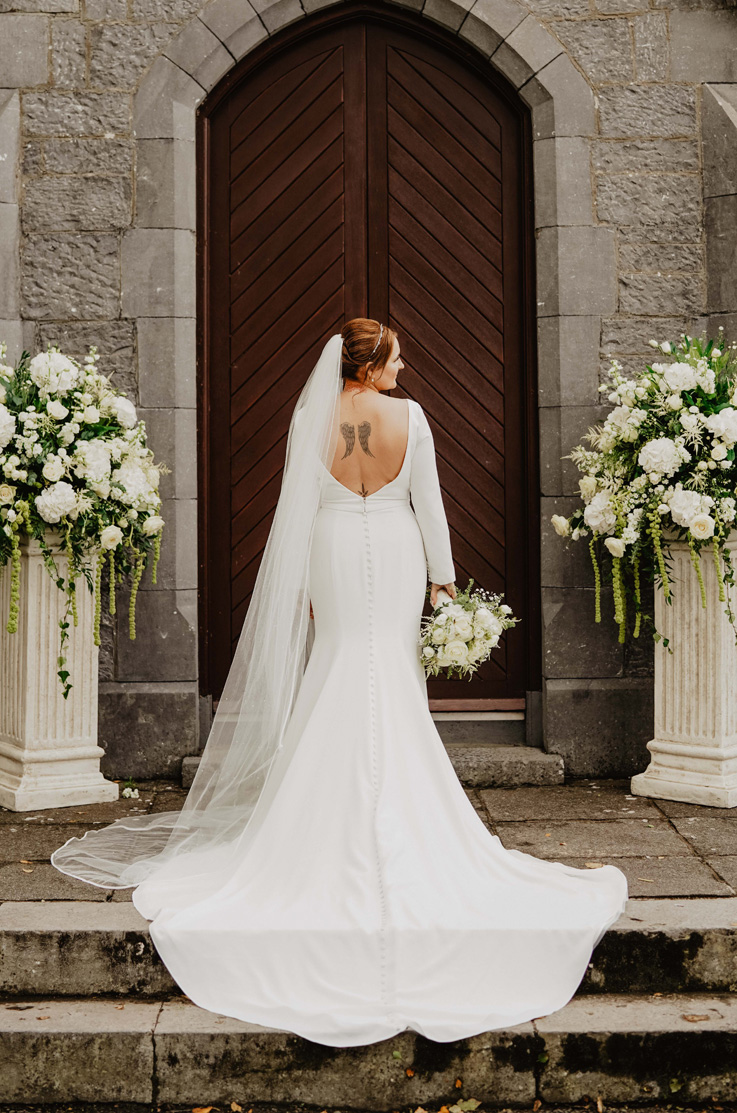 A bride in an open back long sleeve wedding dress and veil, holding a bouquet, stands at the top of stone steps in front of a wooden door with her dress train flowing down the stairs.