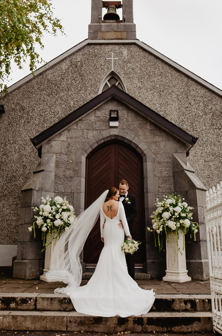 A bride in an open back long sleeve wedding dress and veil, holding a bouquet, and a groom in a suit holding her, stands at he top of stone steps in front of a chapel with a wooden door.