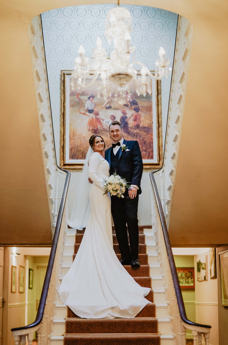 A bride in a long sleeve wedding dress, holding a bouquet, and a groom in a suit, pose on a set of indoor stairs with a chandelier above them and a painting behind them, with the bride's dress train flowing down the stairs.