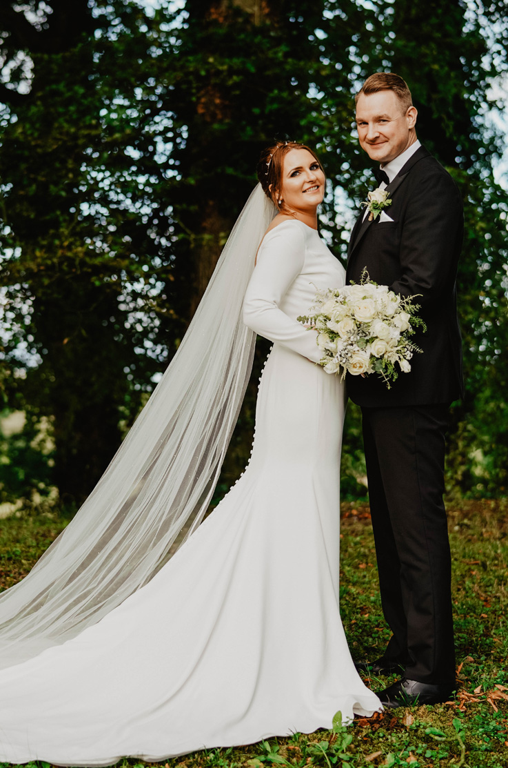 A bride in a long sleeve wedding dress and veil, holding a bouquet, and a groom in a suit, smile and stand together outside with trees in the background.