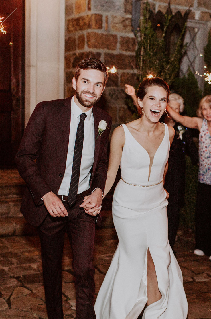 A bride in a skirt slit wedding dress and a groom in a suit hold hands and smile while walking out of a stone building, with wedding guests waving sparklers in the background.