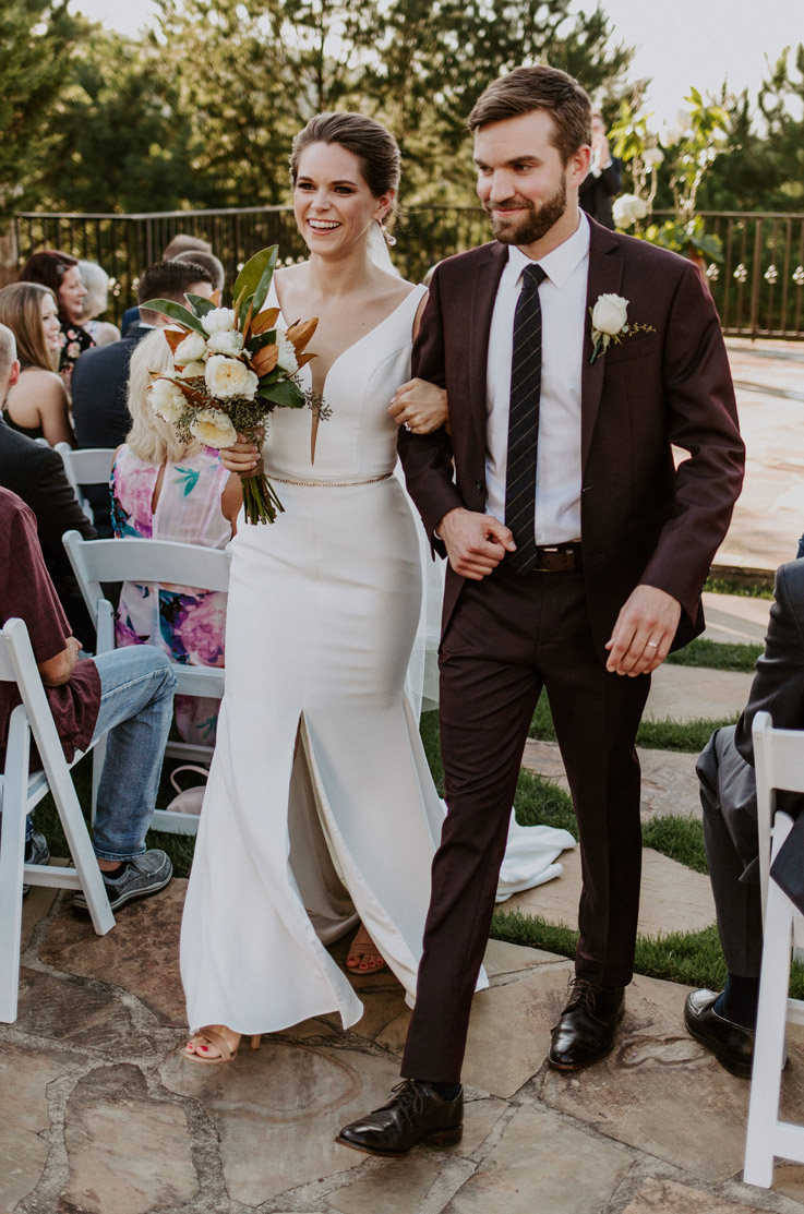 A bride in a skirt slit wedding dress and a groom in a suit walk down an aisle outdoors while smiling and linking arms, with wedding guest watching while seated.