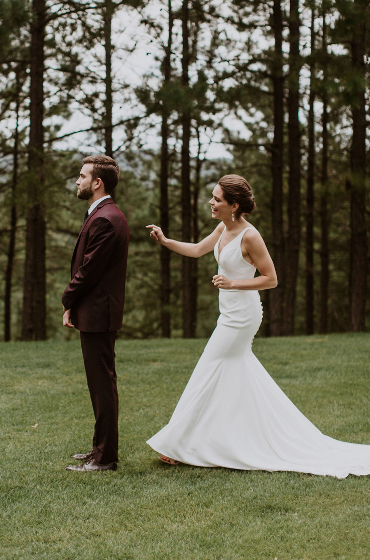 A bride in a sleeveless wedding dress approaches a groom in a suit and is about to tap him on the shoulder as he faces away, both of them outside with trees in the background.