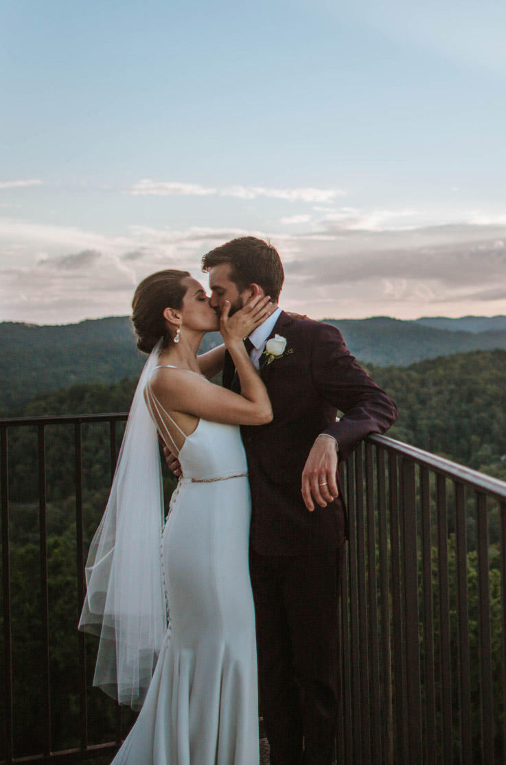 A bride in an embellished wedding dress and veil kisses a groom in a suit as he leans against the railing of an outdoor balcony, with a scenic view of mountains in the background.