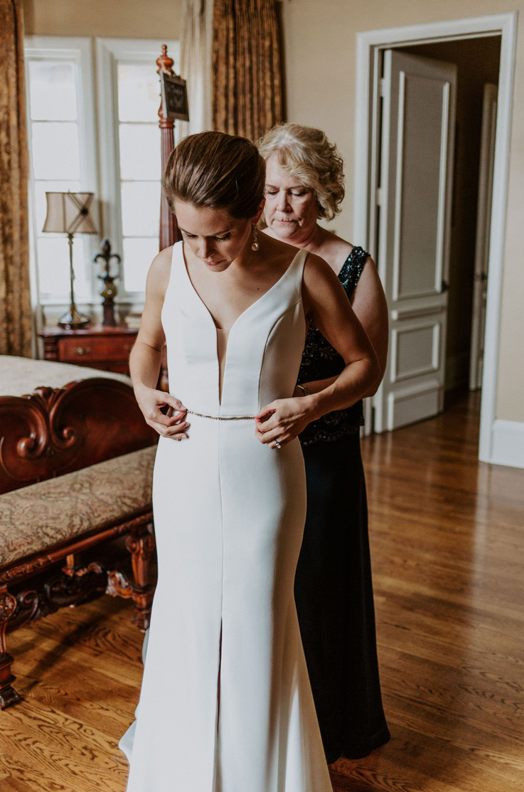 A woman in a sleeveless dress helps a bride in a skirt slit wedding dress fasten her gown as they stand inside a bedroom.