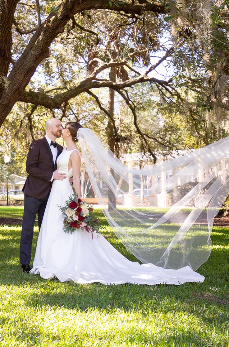 A bride in a sleeveless A-line wedding dress and a veil, holding a bouquet, and a groom in a suit stand outside in the grass and are about to kiss, under tree branches.