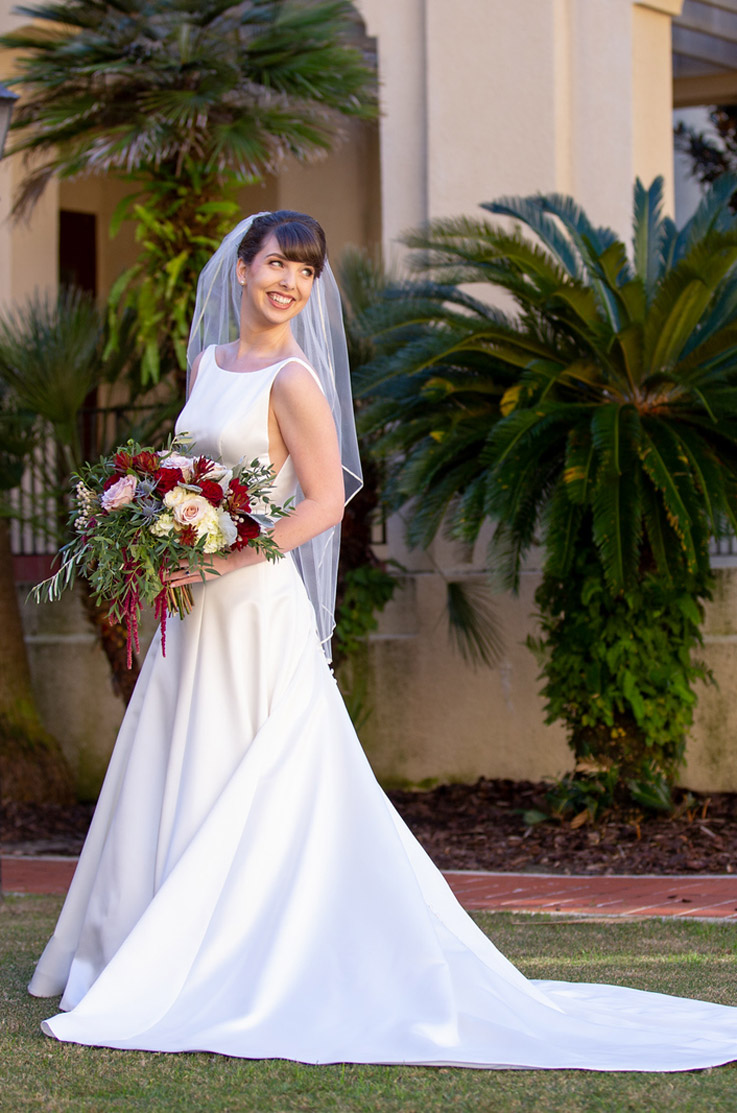A bride in a sleeveless A-line wedding dress and veil, holding a bouquet, smiles and looks over her shoulder as she poses outside in front of a building and short palm trees.
