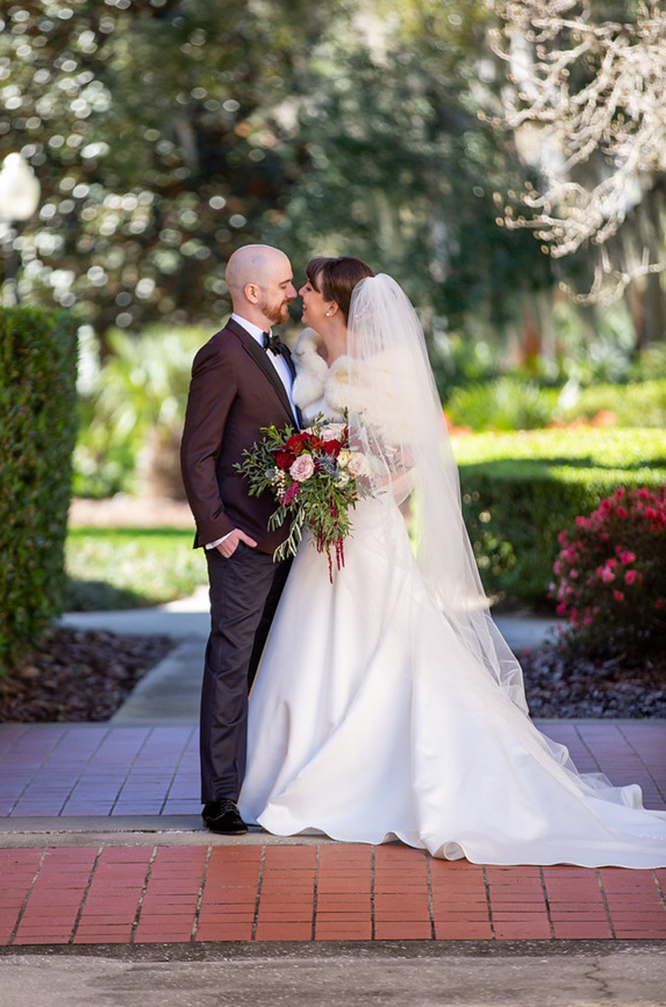 A bride in an A-line wedding dress, a veil, and fur shawl, holding a bouquet, and a groom in a suit, stand outside and are about to kiss, with a garden in the background.