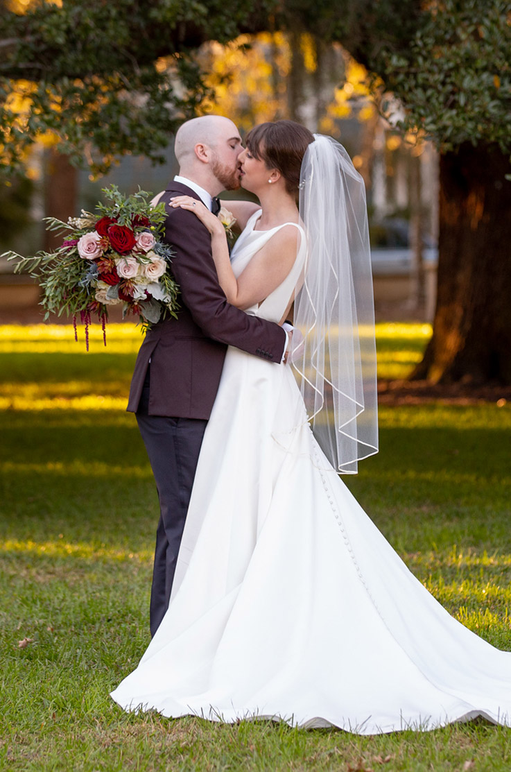 A bride in a sleeveless A-line wedding dress and veil, holding a bouquet, kisses a groom in a suit as they stand outside in the grass with a tree in the background.