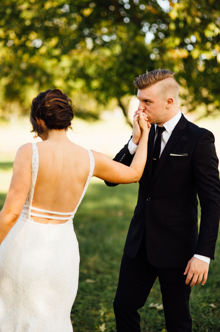 A groom in a suit kisses the hand of a bride in a sleeveless lace wedding dress as they stand outside in the grass, with trees in the background.
