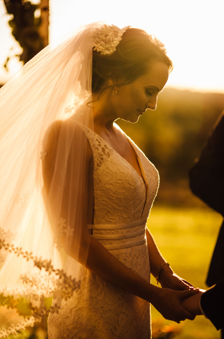A bride in a plunging neckline wedding dress and veil holds the hand of a groom as they stand outside with the sun glowing behind them.