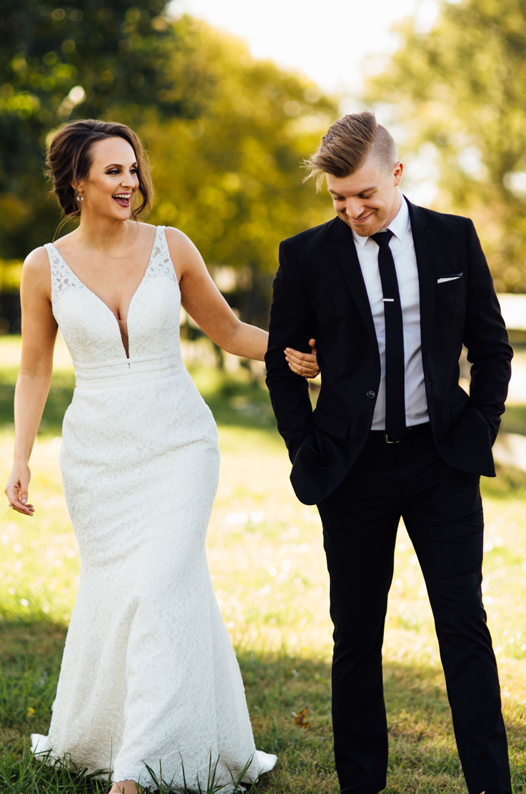 A bride in a plunging neckline lace wedding dress holds the arm of a groom in a suit as they smile and stand outside in the grass, with trees in the background.
