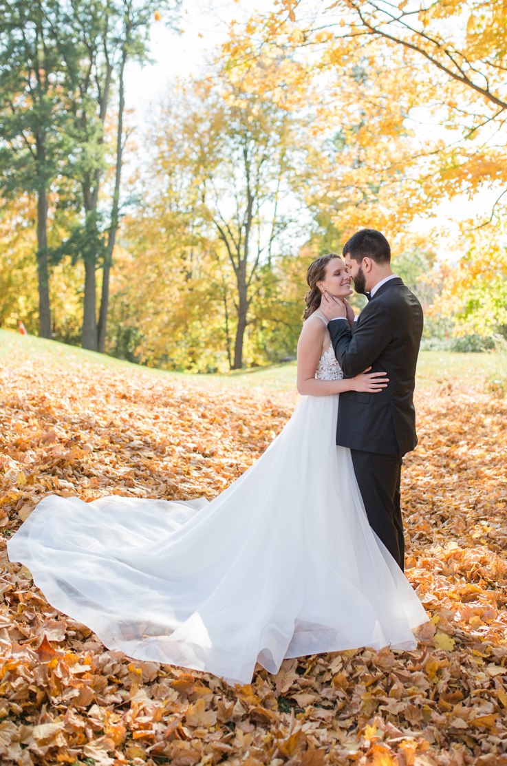 A groom in a suit holds the face of a bride in a lace bodice wedding dress as they stand outside, with fallen leaves beneath them and trees behind them.