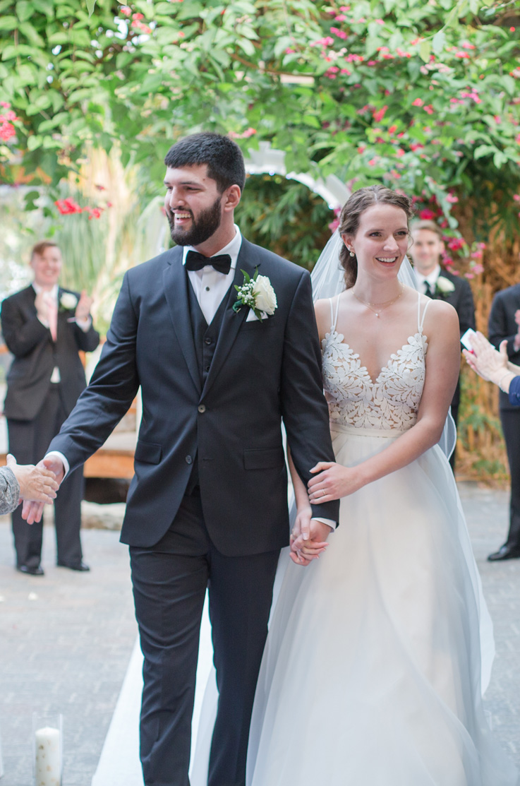 A bride in a lace bodice wedding dress holds the arm of a groom in a suit as they walk down the aisle outside and smile at wedding guests.