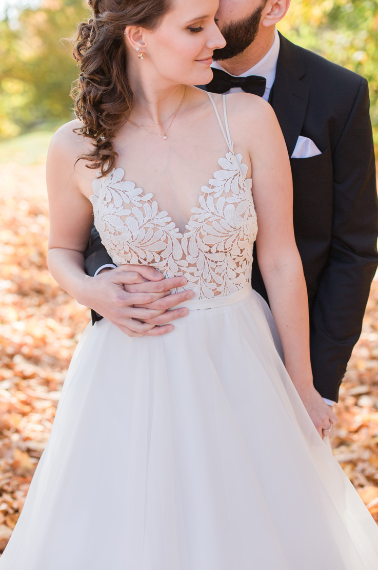 A groom in a suit stands behind a smiling bride in a lace bodice wedding dress and holds her hands while giving her a kiss on the cheek, with fallen leaves beneath them.