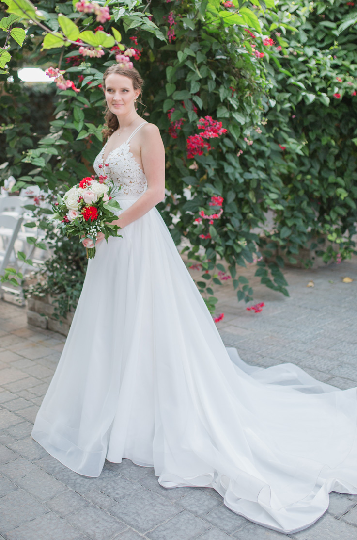 A bride in a lace bodice wedding dress holds a bouquet and poses outside in front of flowering trees, with her dress train flowing behind her. 