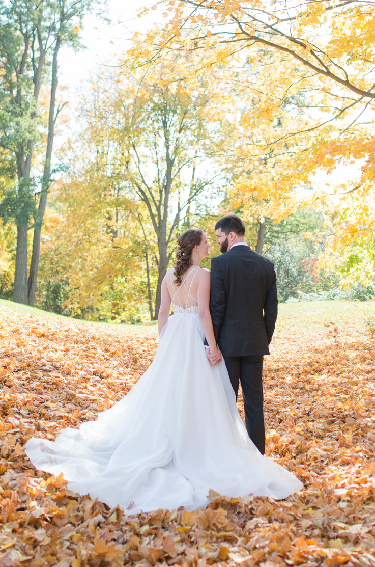 A bride in an Organza wedding dress holds the hand of a groom in a suit as they face away and hold hands, with fallen leaves beneath them and trees behind them.