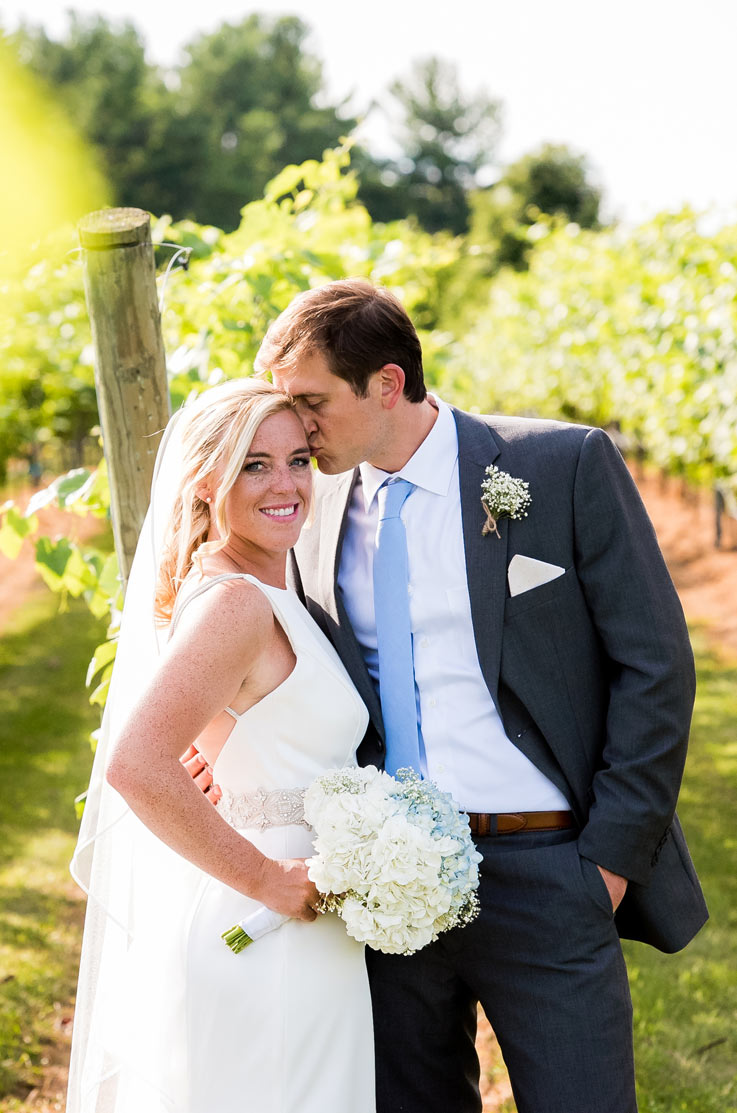 A groom in a suit kisses the temple of a bride in an embellished wedding dress and veil, as she holds a bouquet and smiles, both of them standing outside with a vineyard behind them.