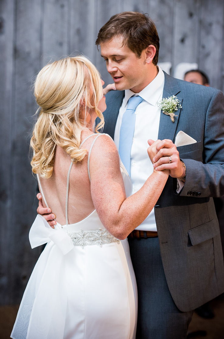 A bride in an embellished wedding dress looks up at a groom in a suit as they dance hand in hand indoors.