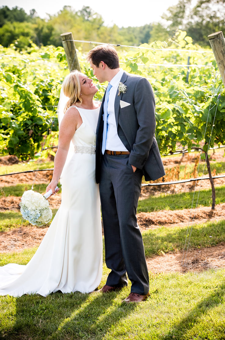 A bride in an embellished wedding dress and veil, holding a bouquet, looks up and smiles at a groom in a suit as he looks at her, both of them standing outside with a vineyard behind them.