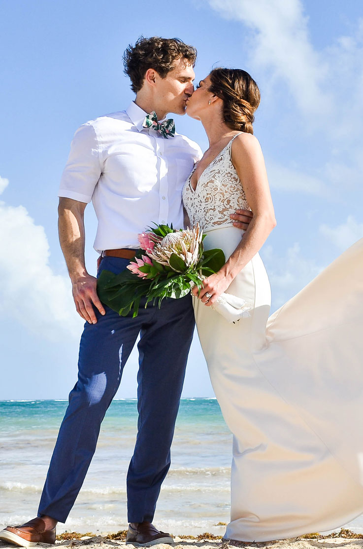 A bride in a lace bodice wedding dress holds a bouquet and kisses a groom in a dress shirt and bow tie as they stand on a sandy beach in front of an ocean.