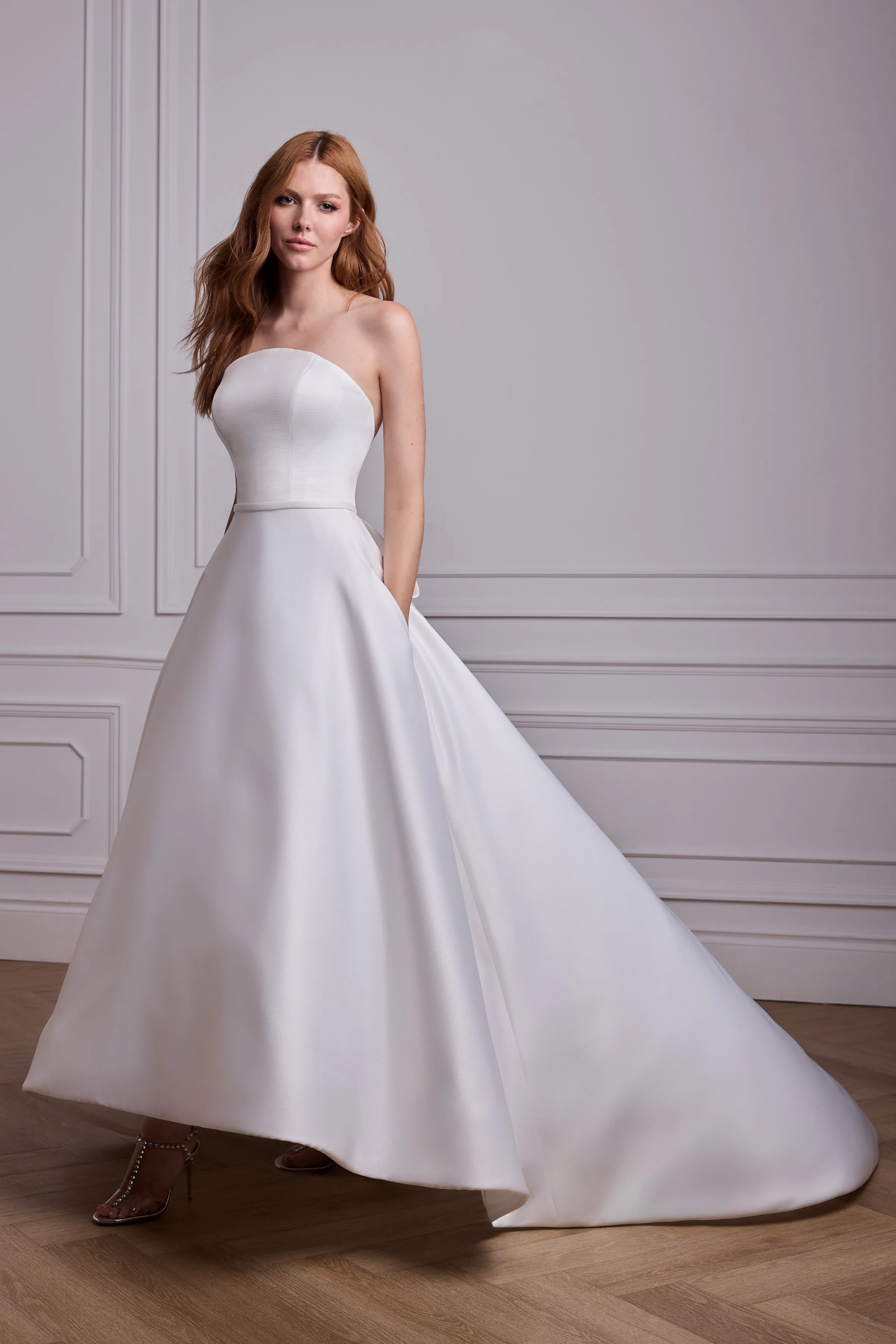 Front view of a strapless Satin Faille arched neckline wedding dress with a band at the waist, and a high-low A-line skirt with pockets, in a ballroom.