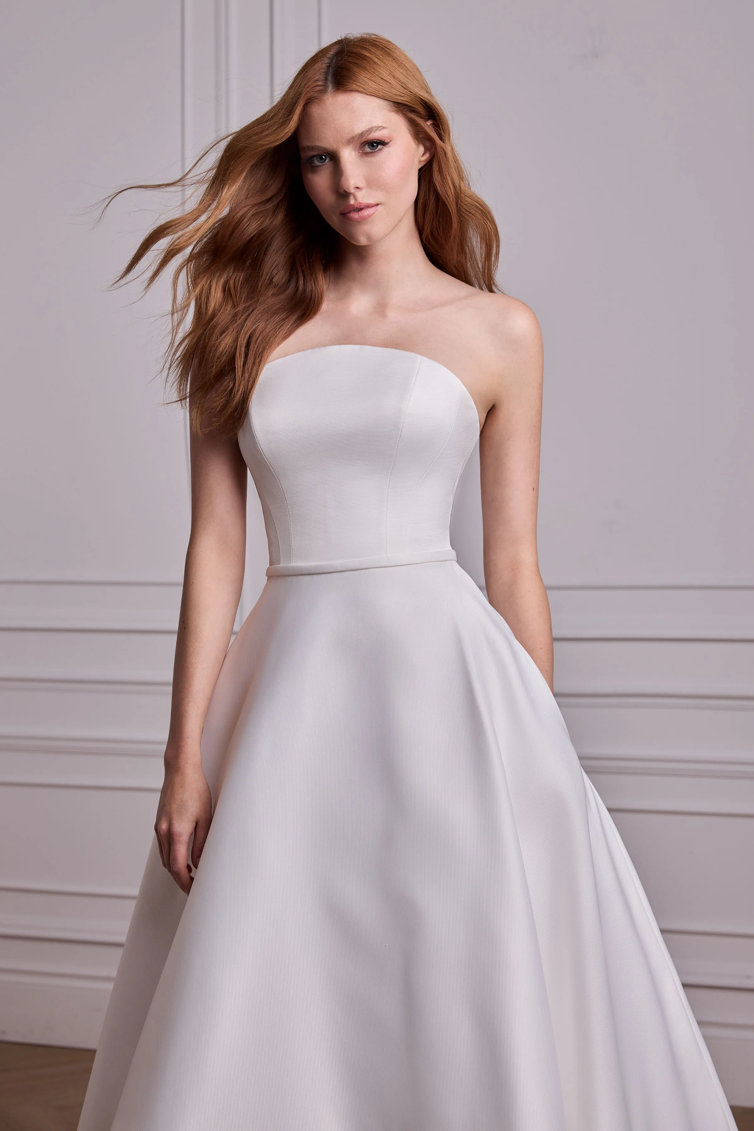 Close up view of a strapless Satin Faille arched neckline wedding dress with a band at the waist, and an A-line skirt, in a ballroom.