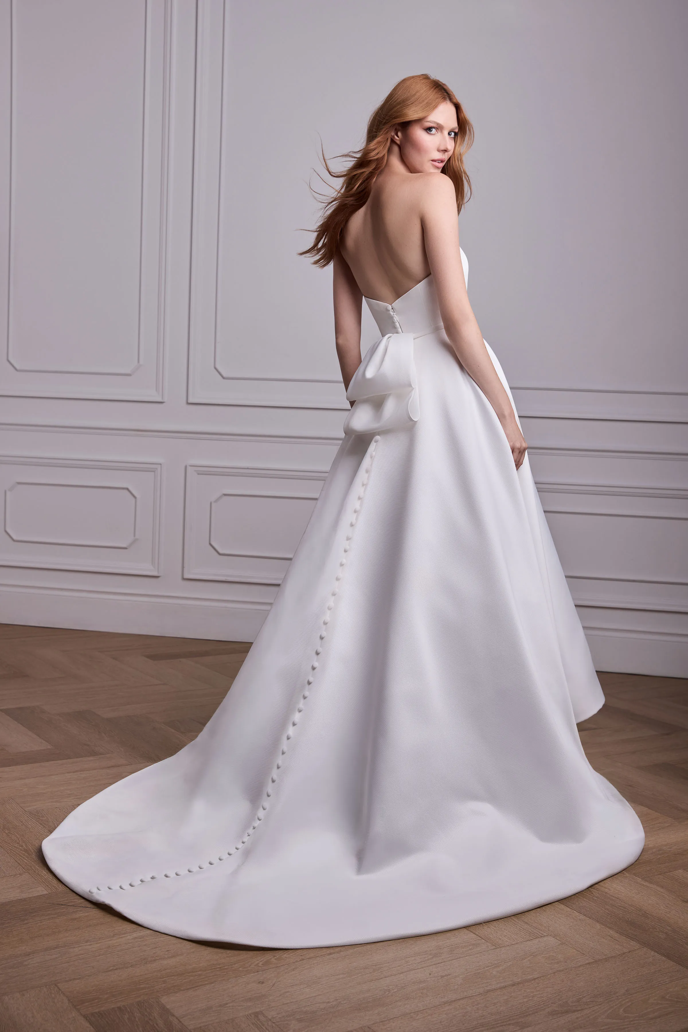 Back view of a strapless Satin Faille wedding dress with a back bow, and buttons down the back of the skirt, in a ballroom.