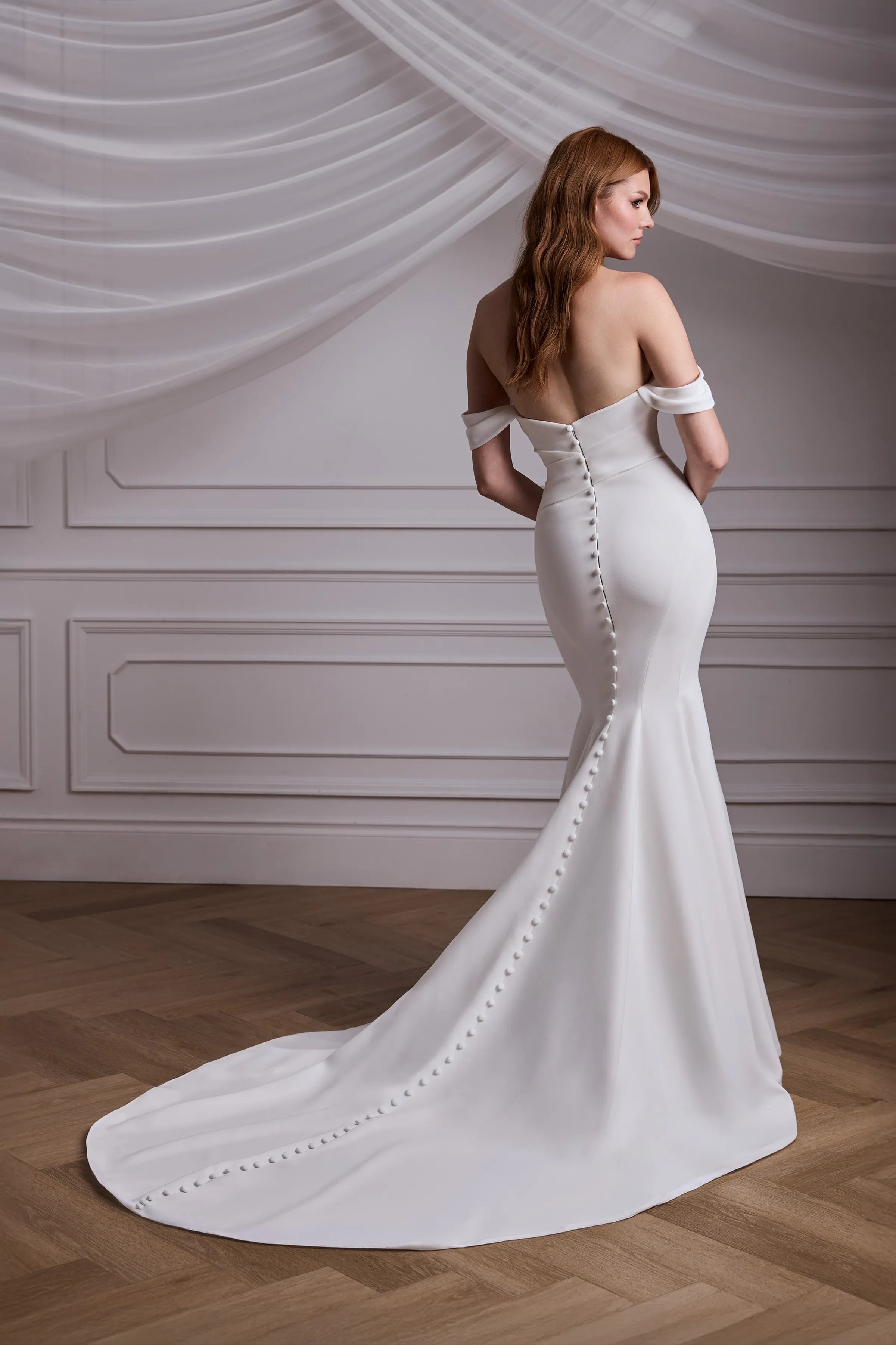 Back view of a strapless fit and flare Crêpe gown with a draped bodice and draped sleeves, with buttons down the back of the gown, in a ballroom with draped tulle in the background.