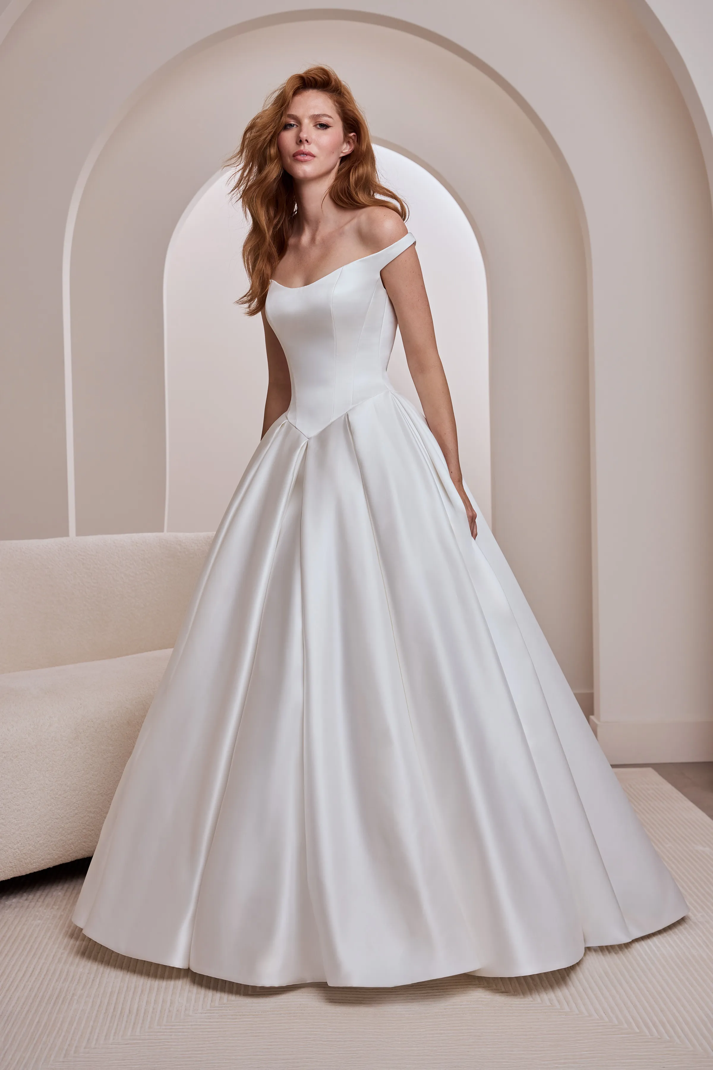 Front view of an off the shoulder Satin wedding dress with a Basque waistline and a pleated ball gown skirt, indoors in front of a couch and an arched wall.