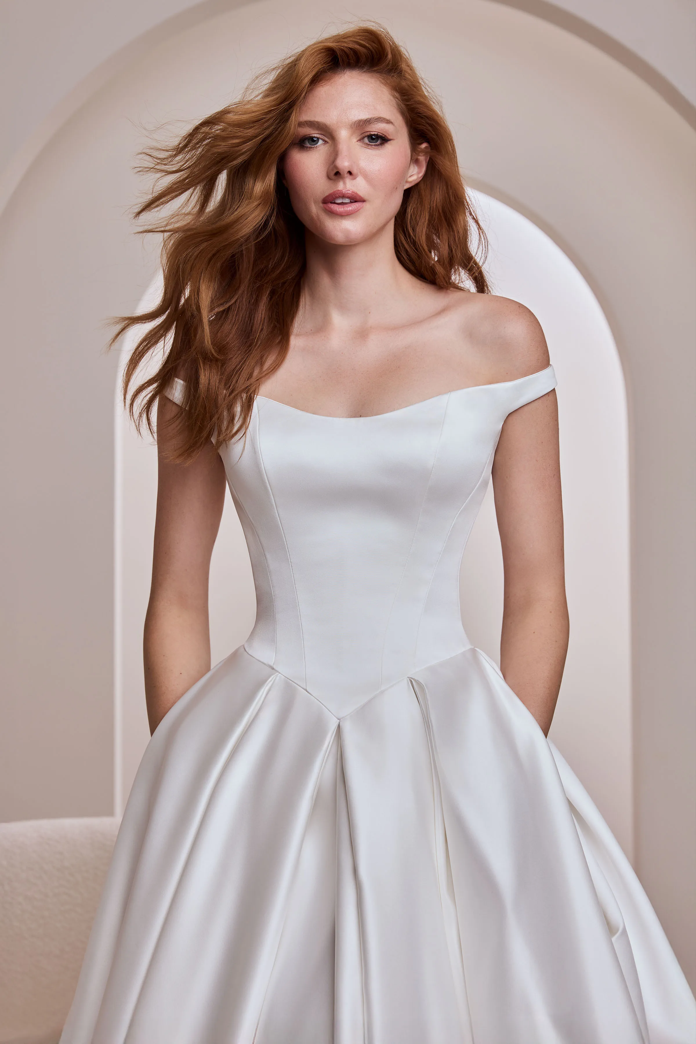 Close up view of an off the shoulder Satin wedding dress with a seamed bodice, Basque waistline, and a pleated skirt with pockets, indoors in front of an arched wall.