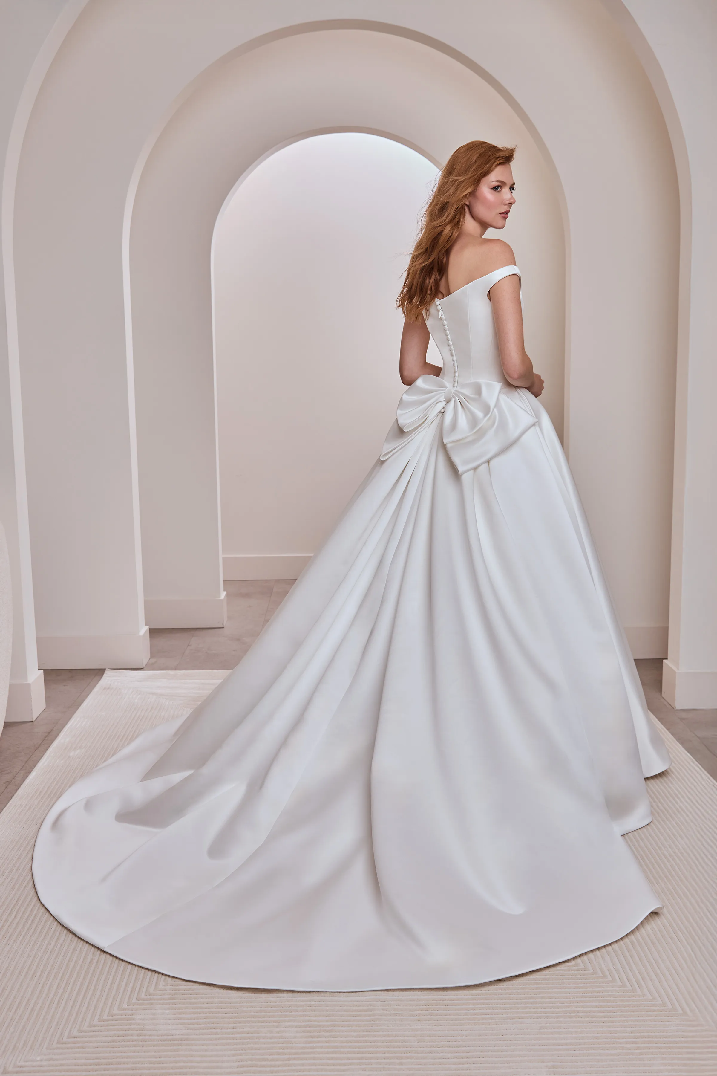 Back view of an off the shoulder Satin wedding dress with buttons down the back of the bodice, a back bow, and a pleated ball gown skirt, indoors in front of an arched wall.