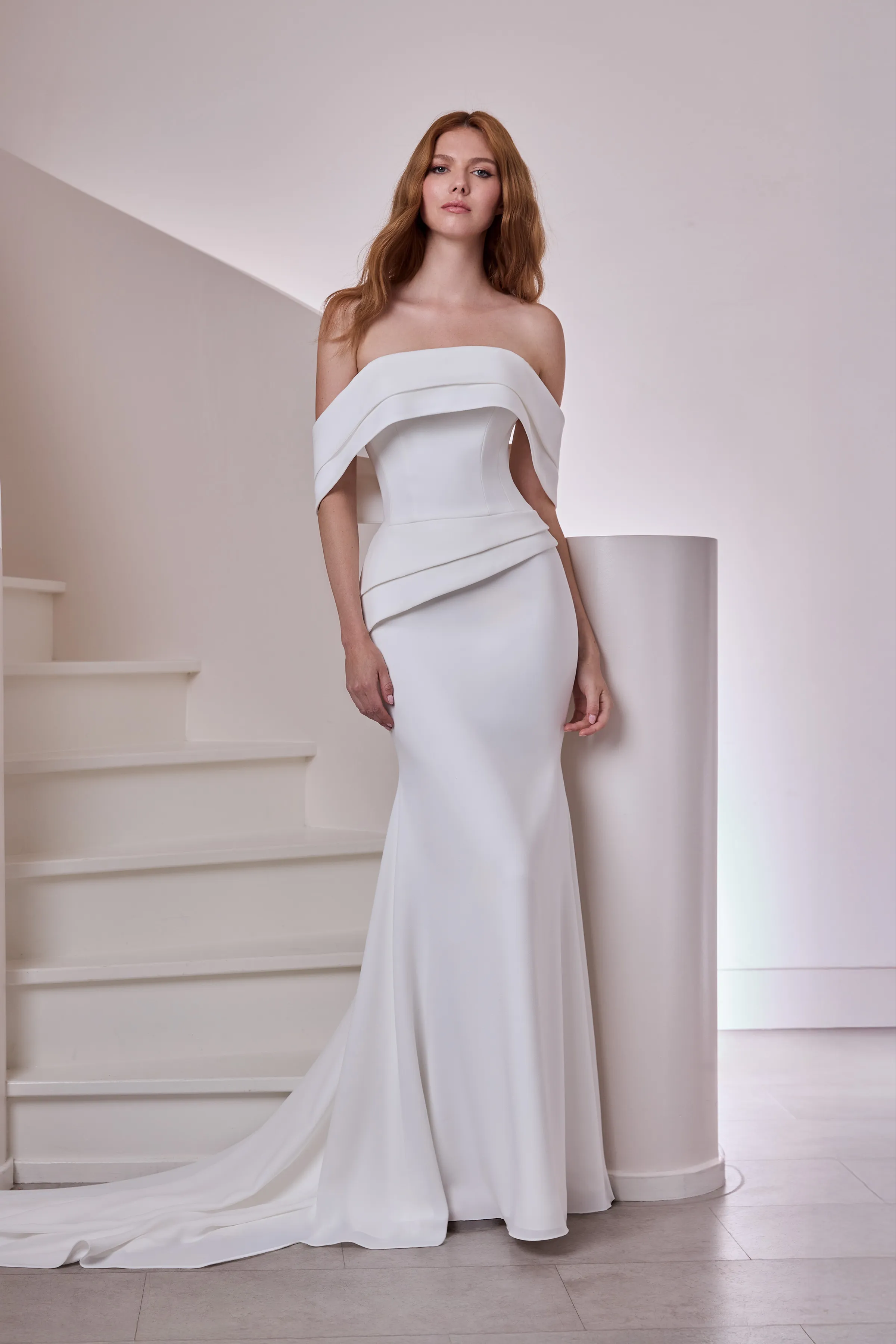 Front view of a strapless straight neckline Crêpe wedding dress with a seamed bodice, an asymmetric peplum waist, and a fit and flare skirt, in front of an indoor spiral staircase.