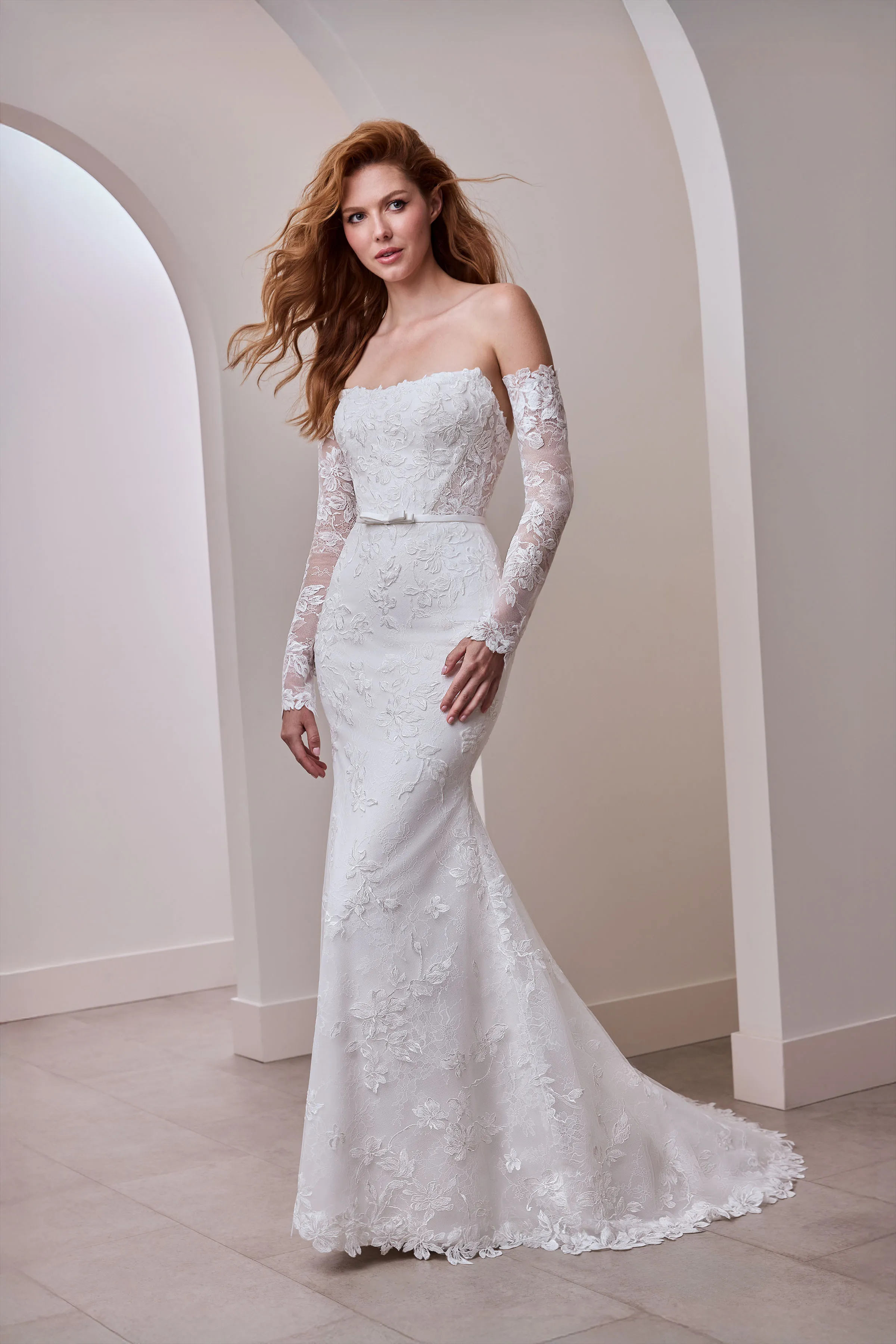 Strapless Lace gown with a scoop neckline, separate long lace sleeves, a band with a bow at the waist, and a lace fit and flare skirt.